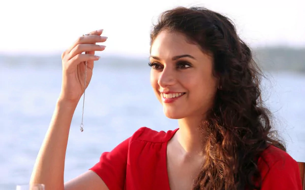 A high-definition desktop wallpaper featuring Bollywood celebrity Aditi Rao Hydari, smiling and holding a necklace, with a scenic water backdrop.