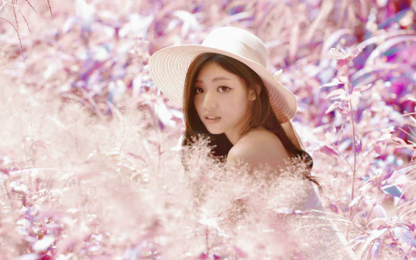 A woman with long hair and a wide-brimmed hat sits in a field of soft pink flowers, surrounded by a dreamy, pastel atmosphere. This stunning image serves as a vibrant 4K Ultra HD desktop wallpaper.