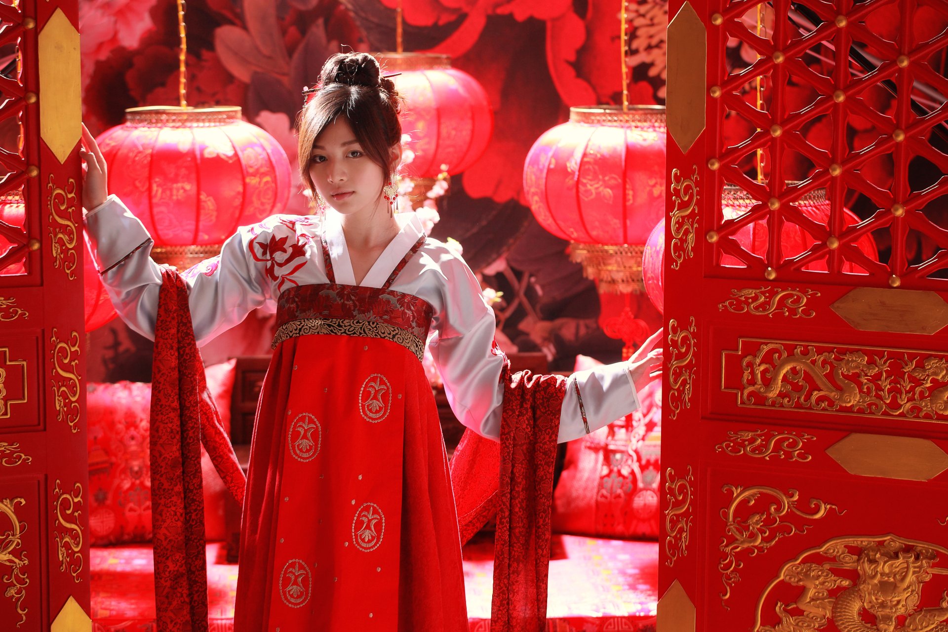 A woman wearing traditional national dress stands gracefully between ornate red doors, surrounded by glowing lanterns, capturing a vibrant celebration of culture.
