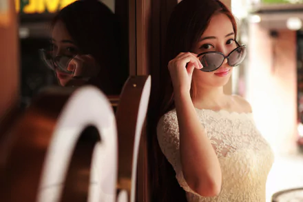 A woman in a white lace dress, holding sunglasses, gazes confidently at the camera. Her reflection is visible in the background.