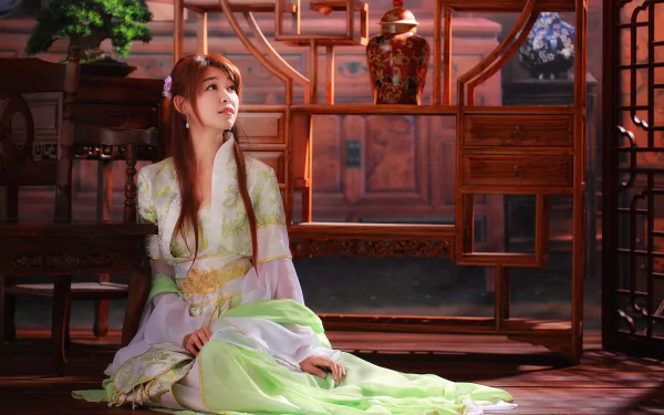 A woman in traditional Asian national dress sits gracefully beside a vase, embodying elegance and serenity in a beautifully lit interior. This HD wallpaper captures a tranquil moment.