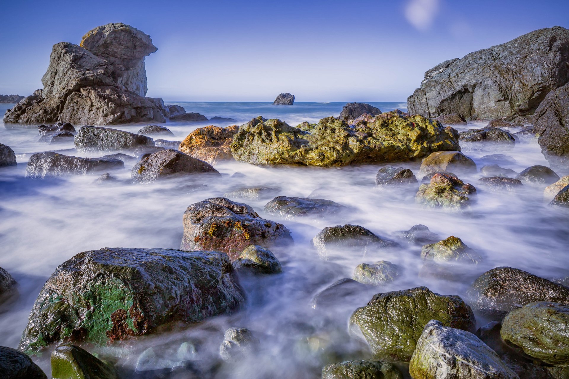 Nature coastline HD PC desktop wallpaper: rocky shore with moss-covered boulders and milky, long-exposure surf under a clear blue sky.