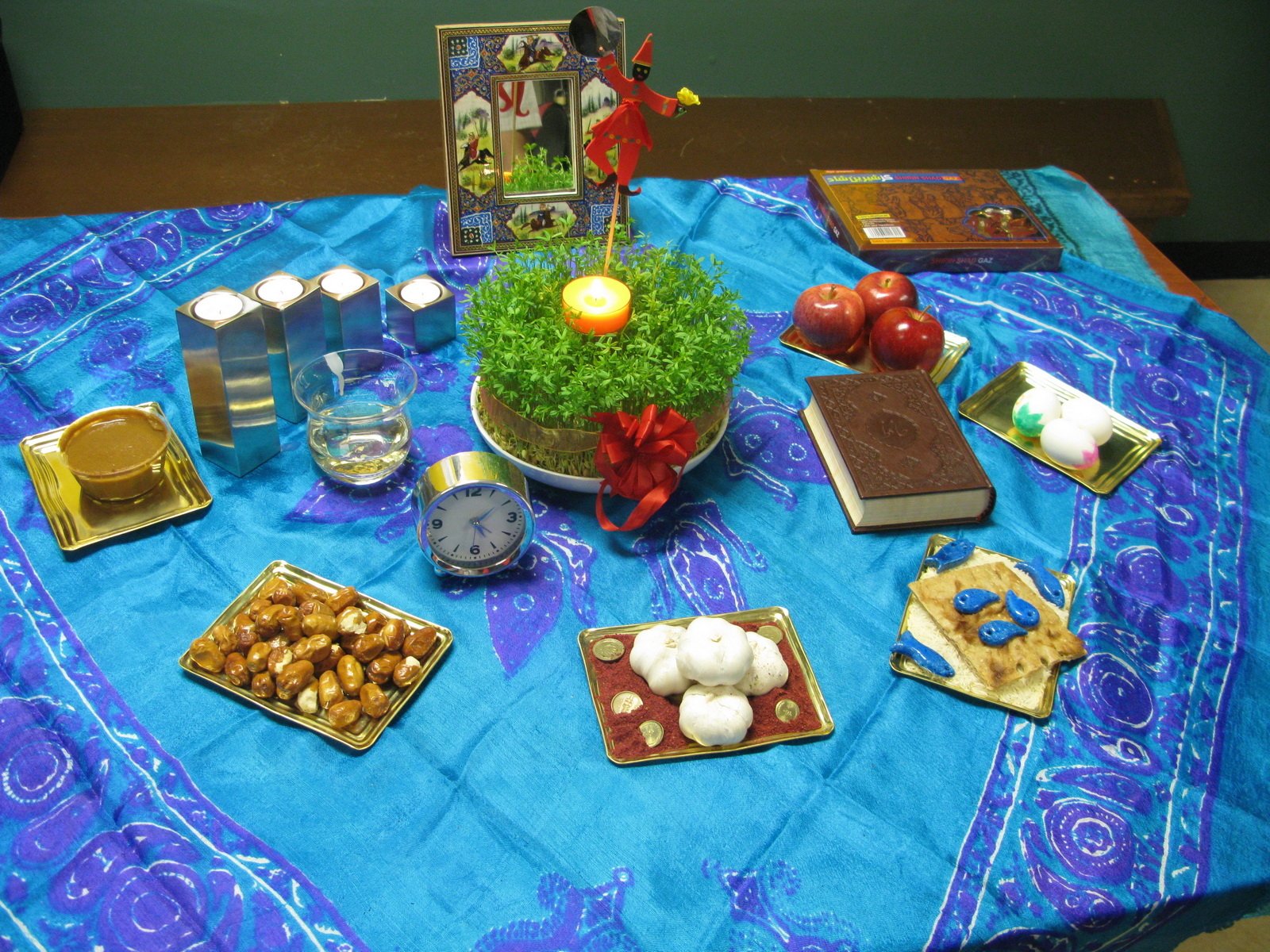 HD PC desktop wallpaper showing a traditional Nowruz holiday table setting with sabzeh, candles, nuts, apples, and other symbolic items on a vibrant blue cloth.