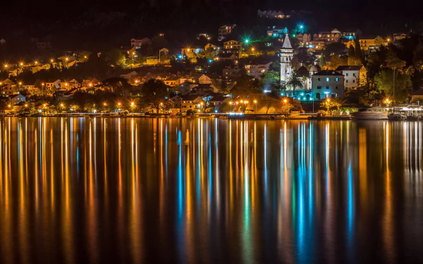 Nighttime HD desktop wallpaper of Kotor, Montenegro: illuminated waterfront and church spires with colorful lights reflected in calm bay waters.
