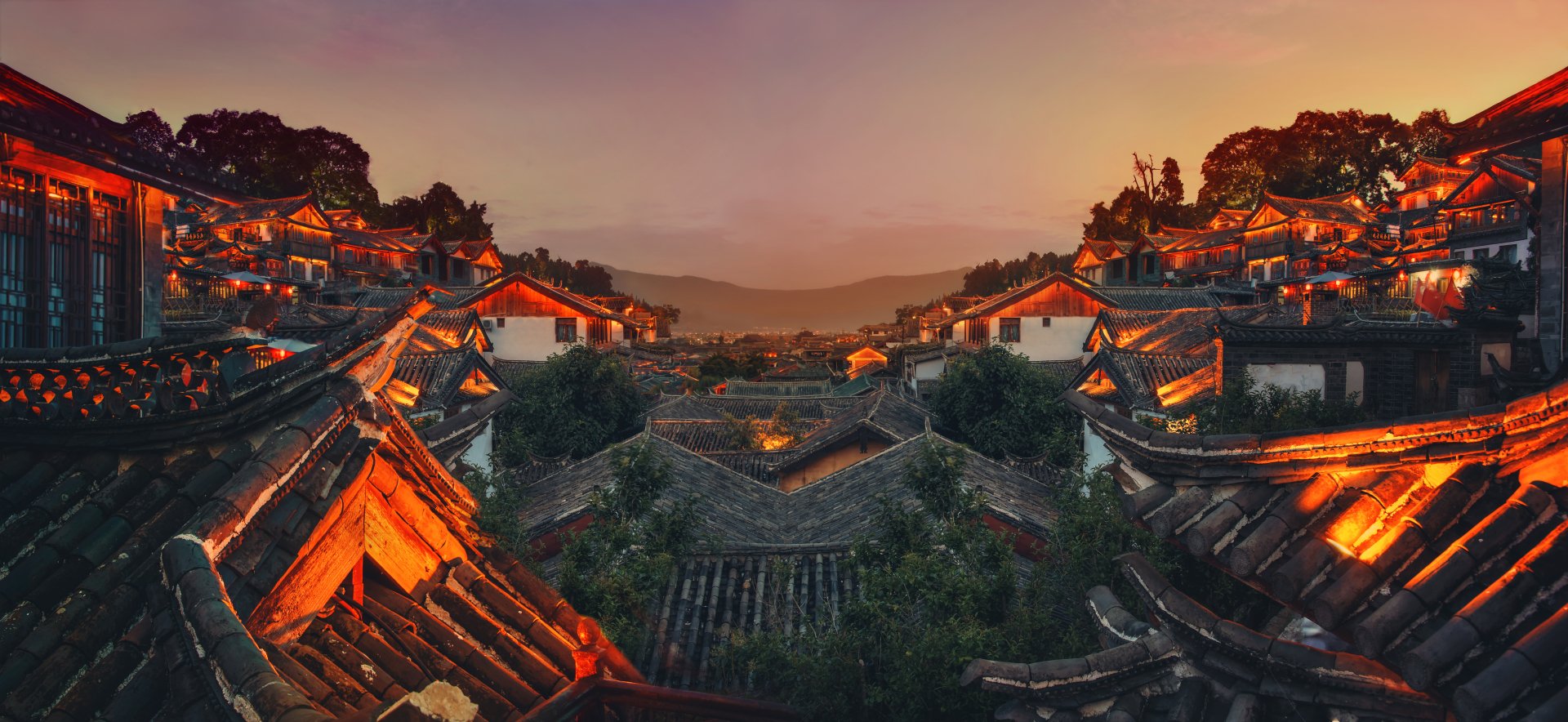 A beautiful HD wallpaper showing the rooftops of a traditional village in Lijiang, Yunnan, China, at sunset, with warm lights illuminating the man-made structures and mountains in the background.