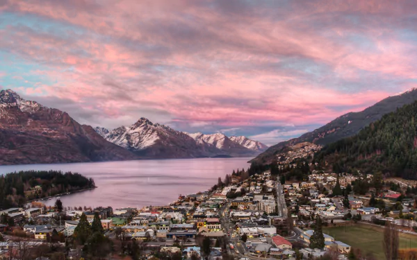 A 4K Ultra HD view of Queenstown, New Zealand, featuring a river, mountain landscape, town, and a pink-hued sky with clouds at sunset.