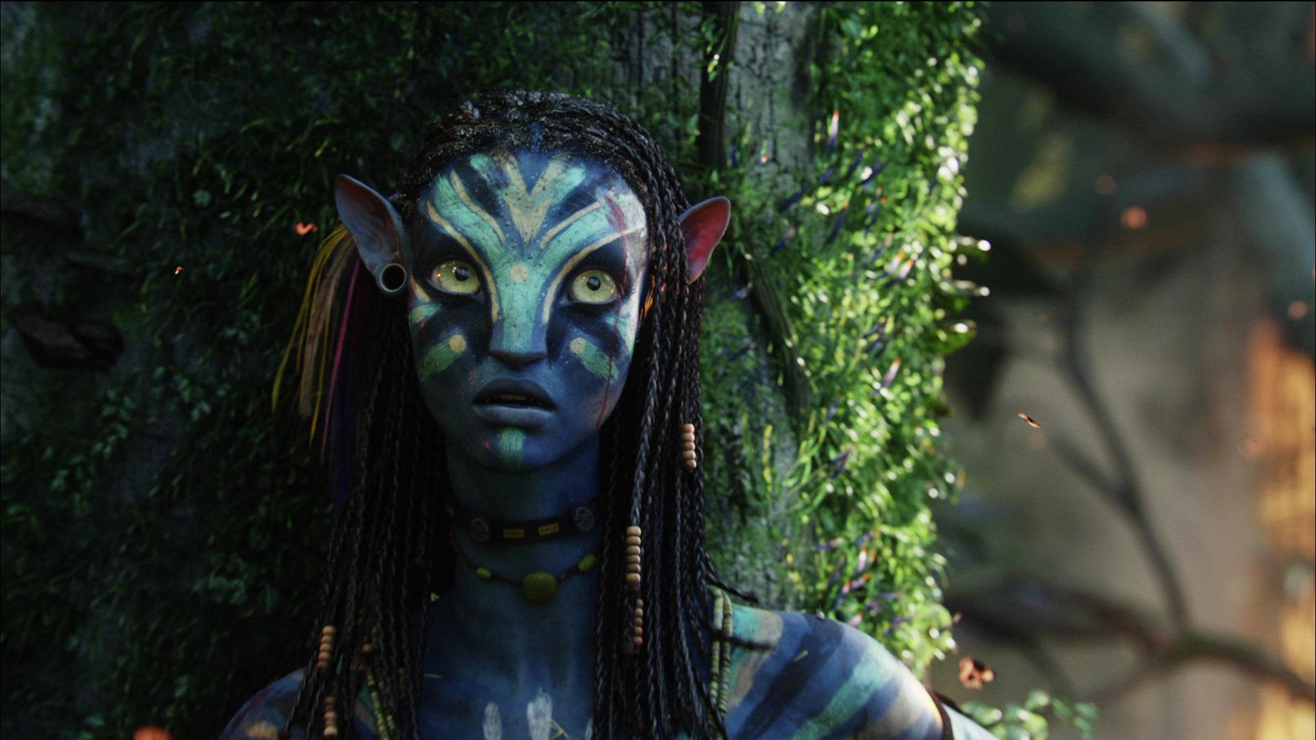 HD PC desktop wallpaper of a blue-skinned Na'vi from the movie Avatar: close-up with braided hair, bioluminescent facial markings and a lush, leafy forest background.