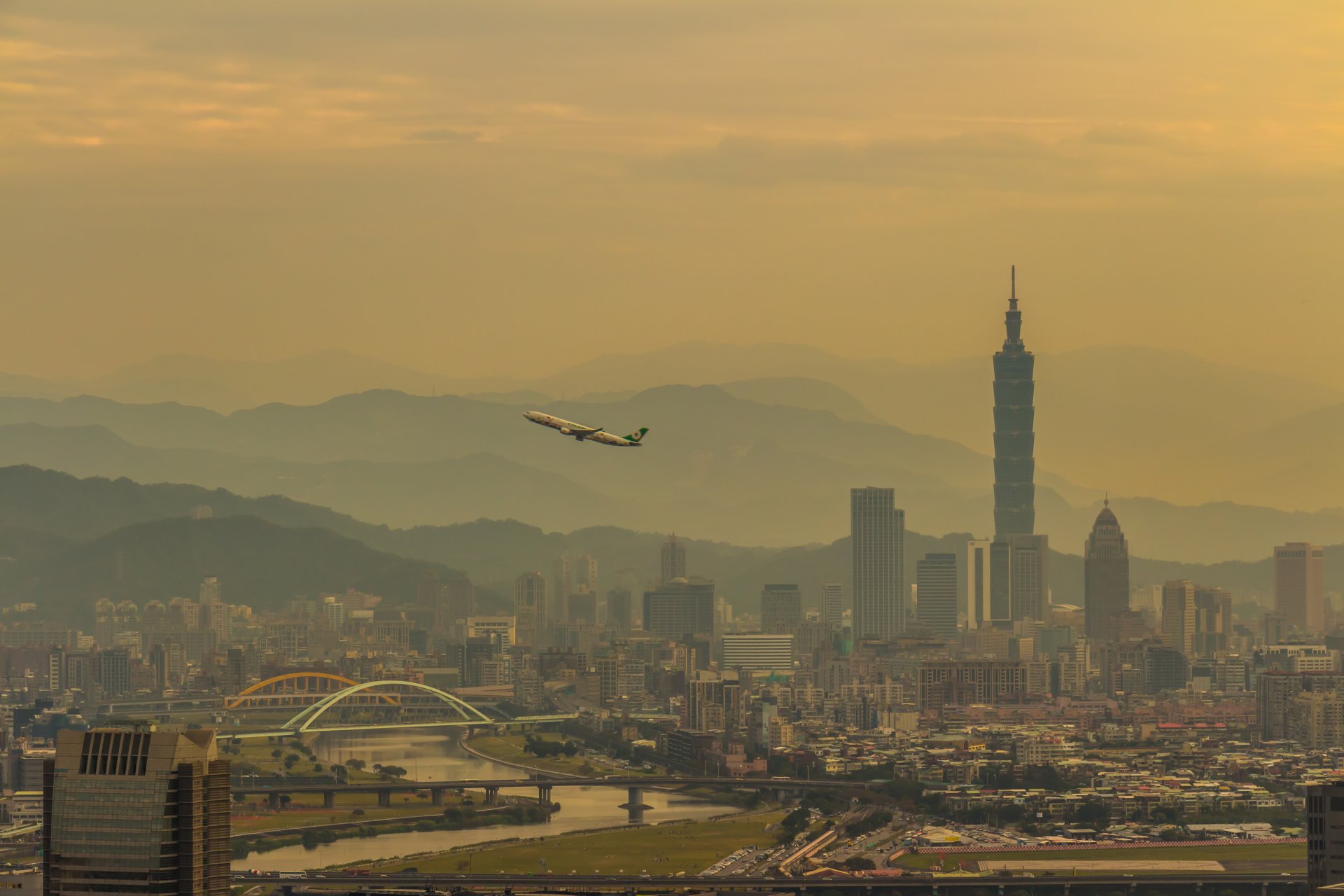 4K Ultra HD wallpaper featuring an airplane flying over Taipei's cityscape with mountains in the background, highlighting Taiwan's blend of man-made and natural beauty.
