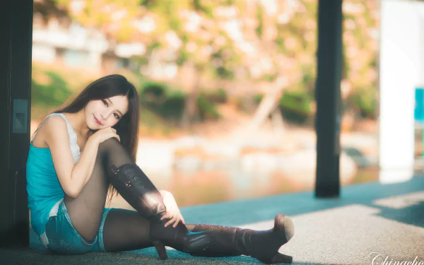 A smiling woman in stylish boots sits gracefully by a riverside, showcasing her relaxed demeanor against a blurred natural backdrop. Taiwan's Zhang Qi Jun captures this moment.