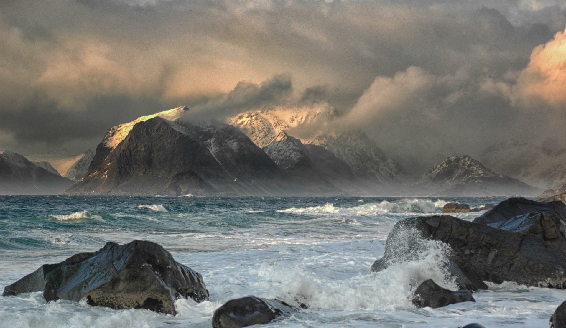 Stormy sea waves crash against rocky shores beneath cloud-covered mountains in Norway's Lofoten Islands, showcasing a dramatic natural seascape.