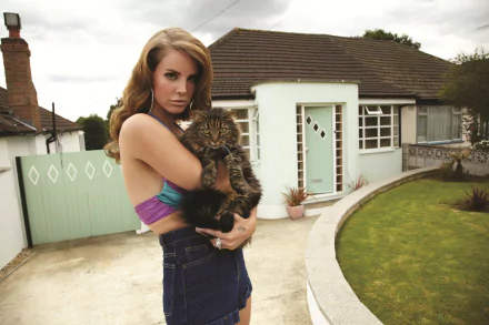 Lana Del Rey holding a fluffy cat outside a suburban house, captured in a 4K Ultra HD desktop wallpaper featuring music and calm outdoor vibes.