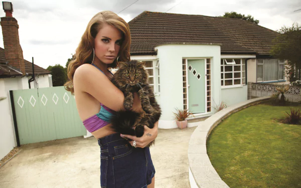 Lana Del Rey holding a fluffy cat outside a suburban house, captured in a 4K Ultra HD desktop wallpaper featuring music and calm outdoor vibes.