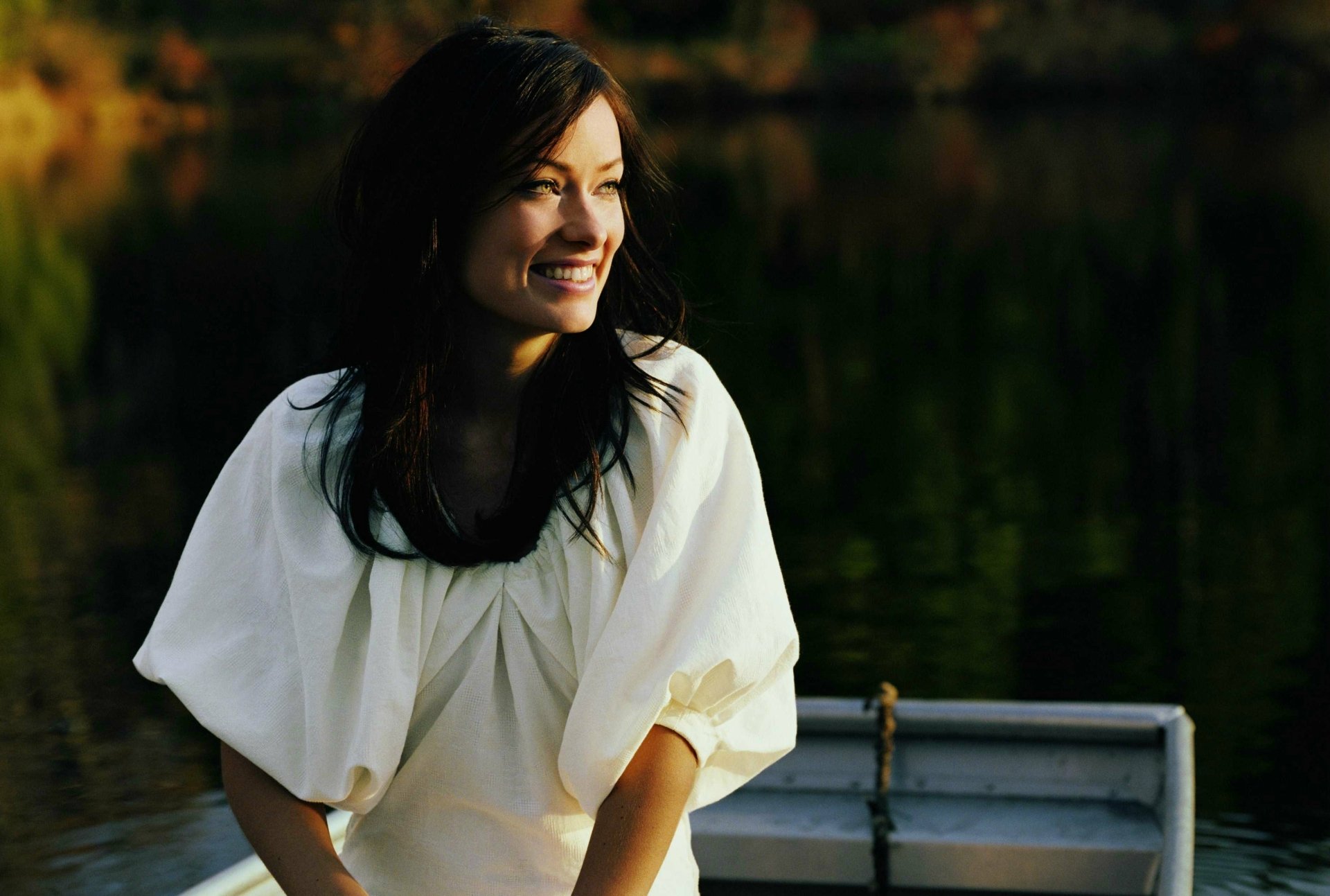 HD desktop wallpaper featuring celebrity Olivia Wilde smiling warmly while sitting in a boat on calm water with a dark, natural background.