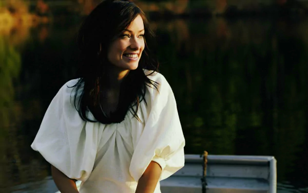 HD desktop wallpaper featuring celebrity Olivia Wilde smiling warmly while sitting in a boat on calm water with a dark, natural background.
