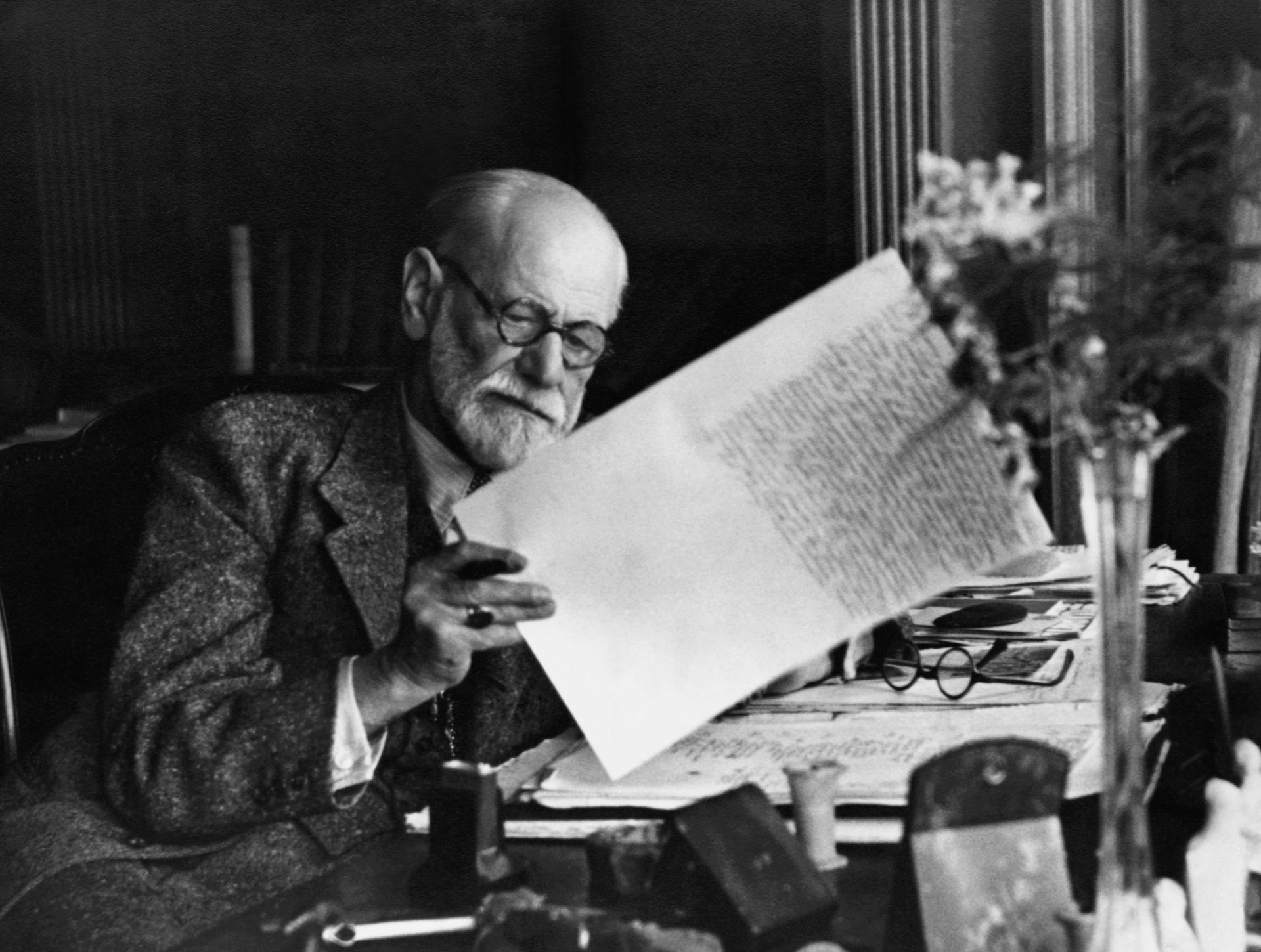 Black and white 4K Ultra HD wallpaper of Sigmund Freud, an elderly man with glasses, carefully examining a handwritten document at his desk.
