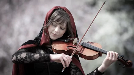HD desktop wallpaper featuring Lindsey Stirling playing the violin, dressed in a dark lace outfit with a red hood, set against a blurred natural background.