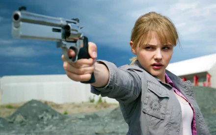 Chloë Grace Moretz from the movie Kick-Ass 2 points a revolver forward, set against a dramatic outdoor backdrop. HD desktop wallpaper and background.