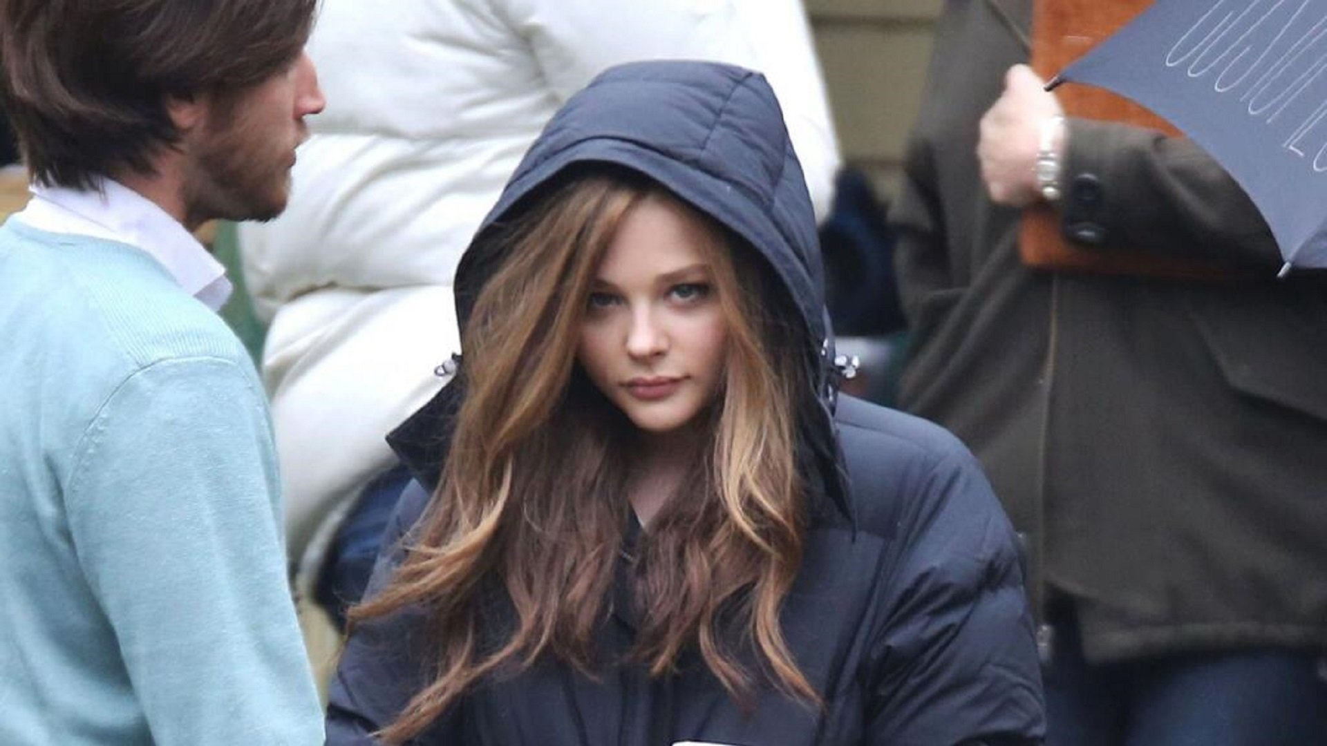 A high-definition desktop wallpaper featuring celebrity Chloë Grace Moretz, dressed in a heavy coat with the hood up, amidst a casual outdoor setting with other people nearby.