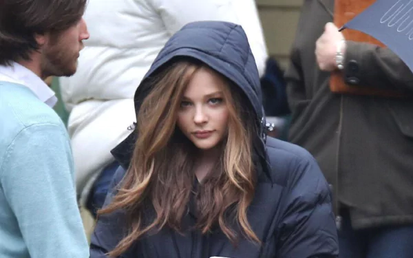 A high-definition desktop wallpaper featuring celebrity Chloë Grace Moretz, dressed in a heavy coat with the hood up, amidst a casual outdoor setting with other people nearby.