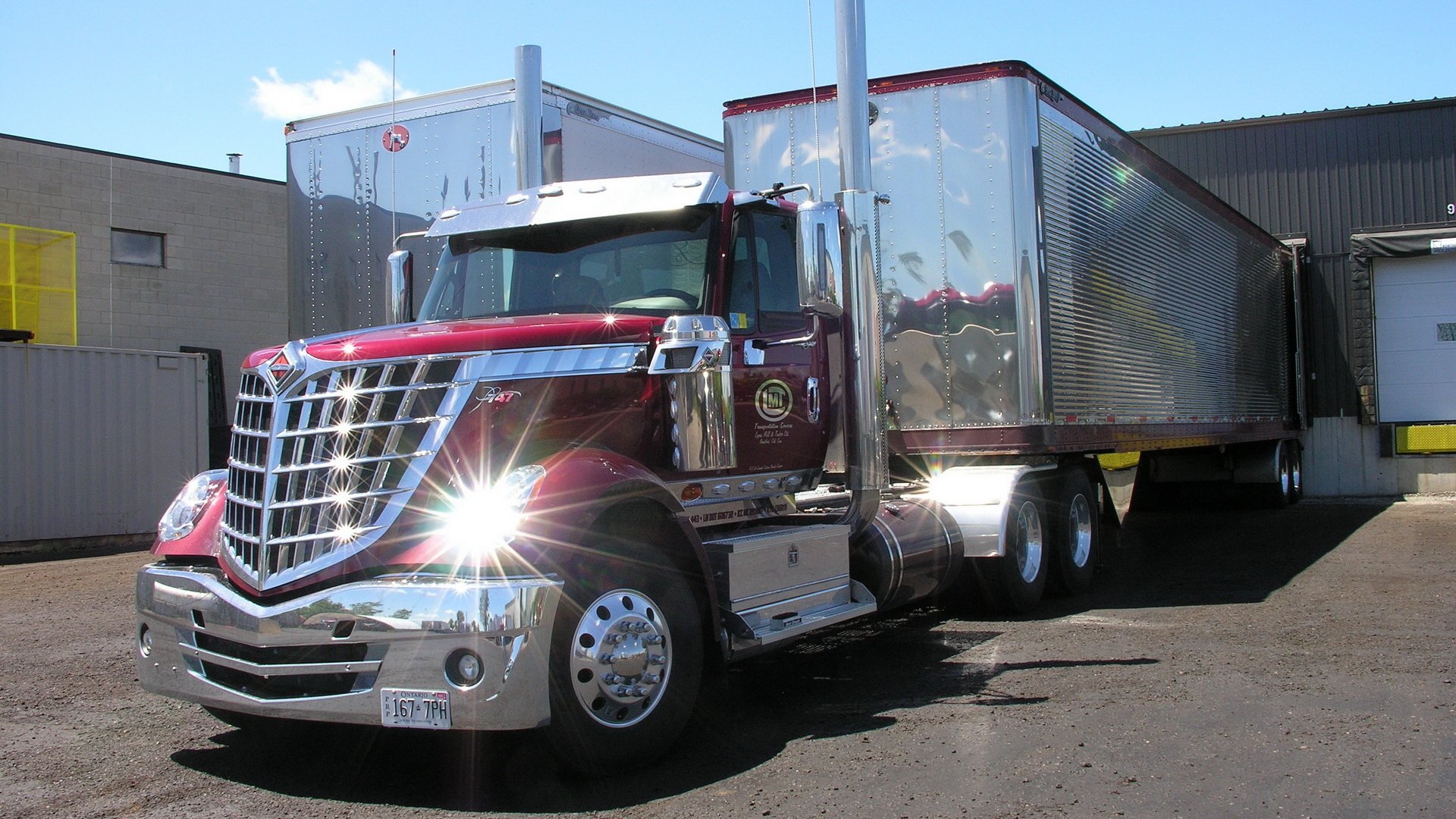 A vibrant HD desktop wallpaper featuring a shiny red truck parked beside a metallic trailer, showcasing its impressive design and polished chrome details against a clear blue sky.