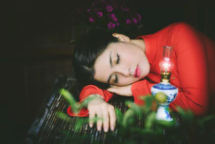 A serene Vietnamese woman in a red dress rests beside a vintage lamp, with vibrant flowers in the background, creating a tranquil HD wallpaper scene.
