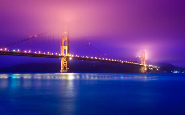 4K Ultra HD desktop wallpaper of the Golden Gate Bridge illuminated at night, with glowing lights reflecting on the calm water under a purple-hued sky.