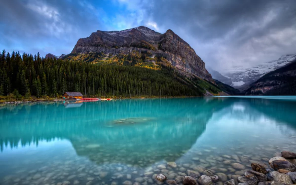 HD desktop wallpaper showcasing the serene turquoise waters of Lake Louise surrounded by dense forest and majestic mountain peaks under a cloudy sky.