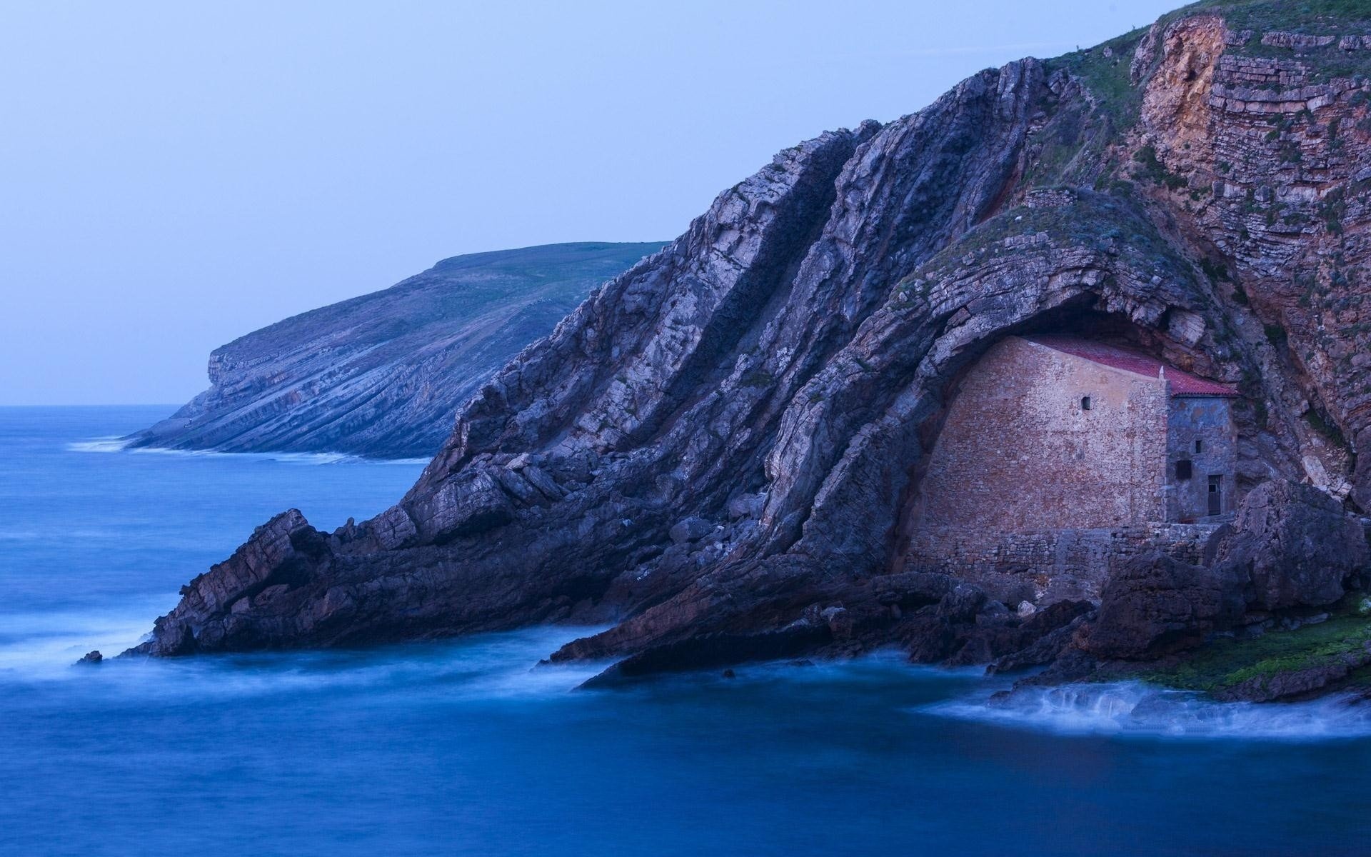 A man-made stone house built into a rocky cliff by the ocean, captured in a serene HD PC desktop wallpaper showcasing natural and architectural contrast.