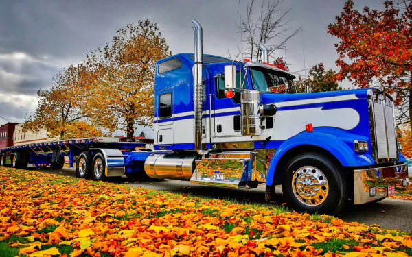 A vibrant blue Kenworth truck parked amid colorful fall foliage, showcasing the beauty of autumn with fallen leaves scattered along the roadside. High-definition desktop wallpaper.