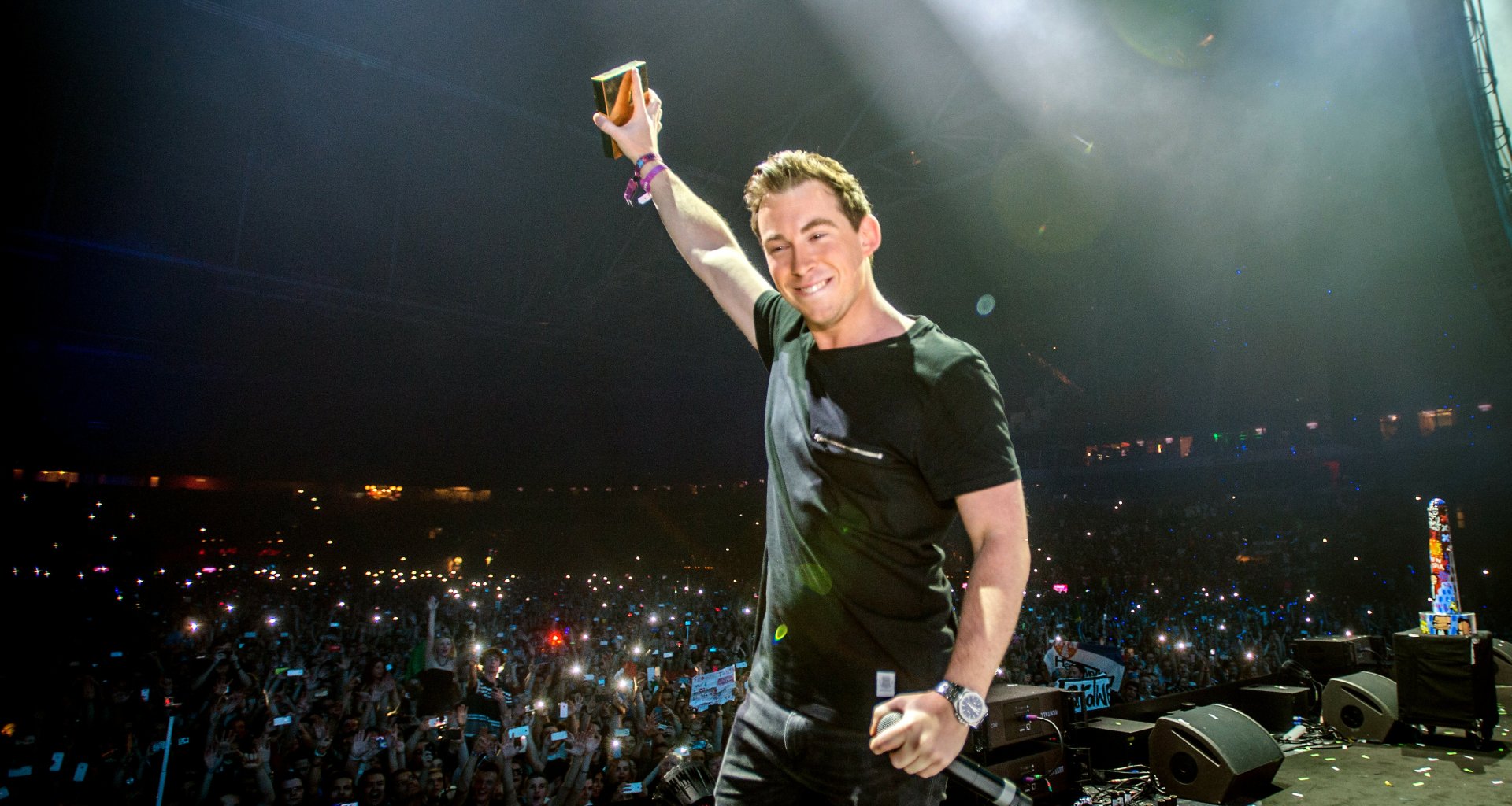 A high-definition wallpaper featuring DJ Hardwell joyfully raising a cup on stage, surrounded by an enthusiastic crowd illuminated by lights, capturing the energy of his performance.