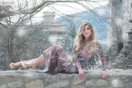 A blonde woman in a dress and high heels poses on a stone wall outdoors during snowfall. The serene setting creates a captivating mood, captured in this HD desktop wallpaper and background.