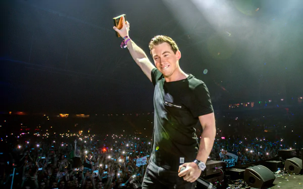 A high-definition wallpaper featuring DJ Hardwell joyfully raising a cup on stage, surrounded by an enthusiastic crowd illuminated by lights, capturing the energy of his performance.