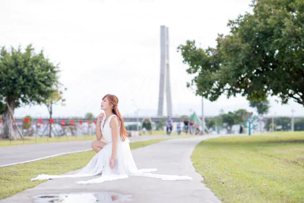 Woman model in a flowing white dress kneels on a park path beside trees with a bridge in the background — 5K Ultra HD PC desktop wallpaper/background.