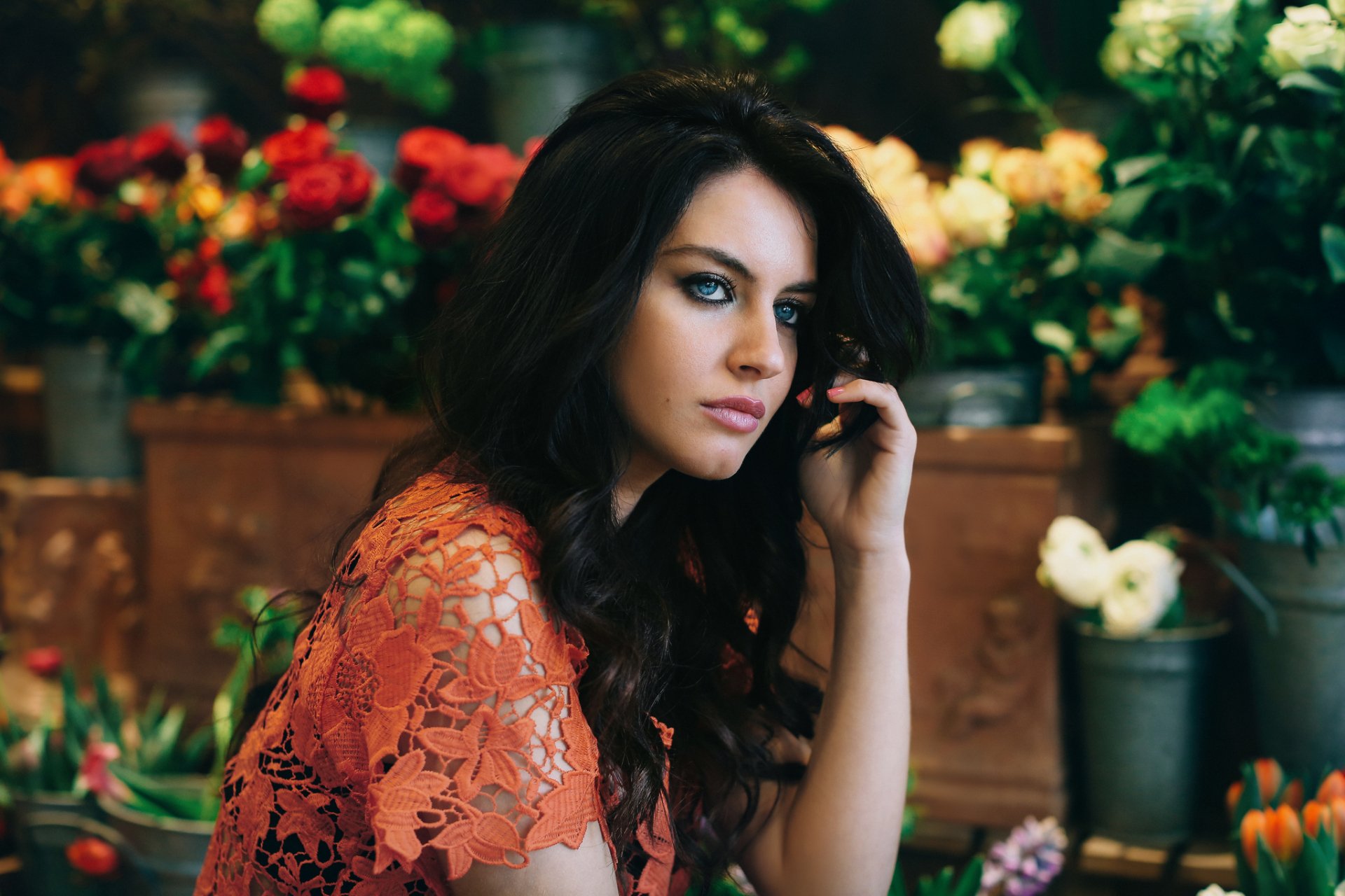 A HD desktop wallpaper of a woman model with black hair and blue eyes, wearing a lace top, sitting among vibrant flowers.