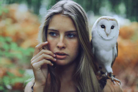 A blonde woman with blue eyes gazes thoughtfully, her hand touching her face, as a barn owl perches on her shoulder, surrounded by a lush forest backdrop.
