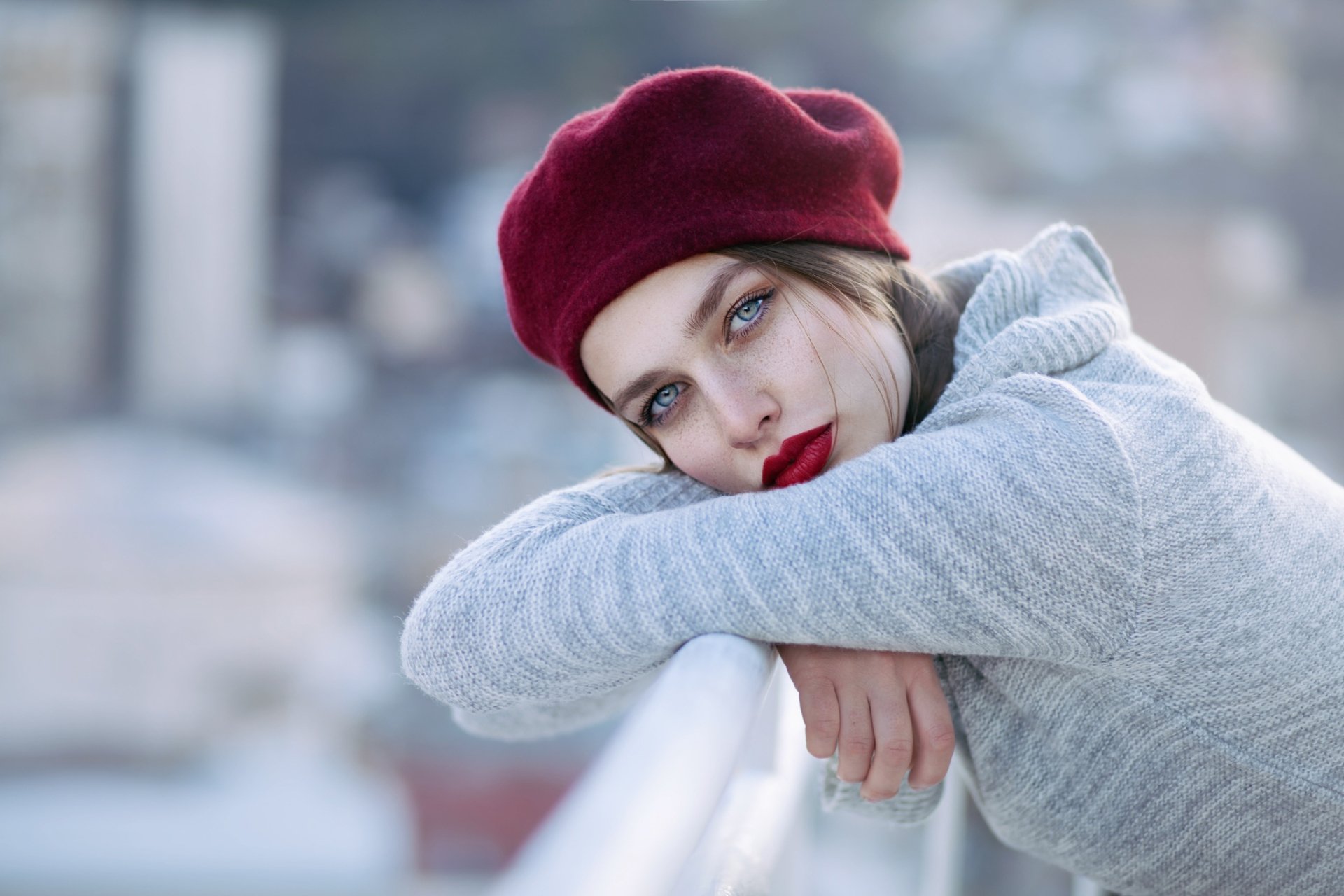 A brunette woman with striking green eyes leans on a railing, wearing a red beret and a gray sweater, surrounded by a soft bokeh background, capturing a serene moment.