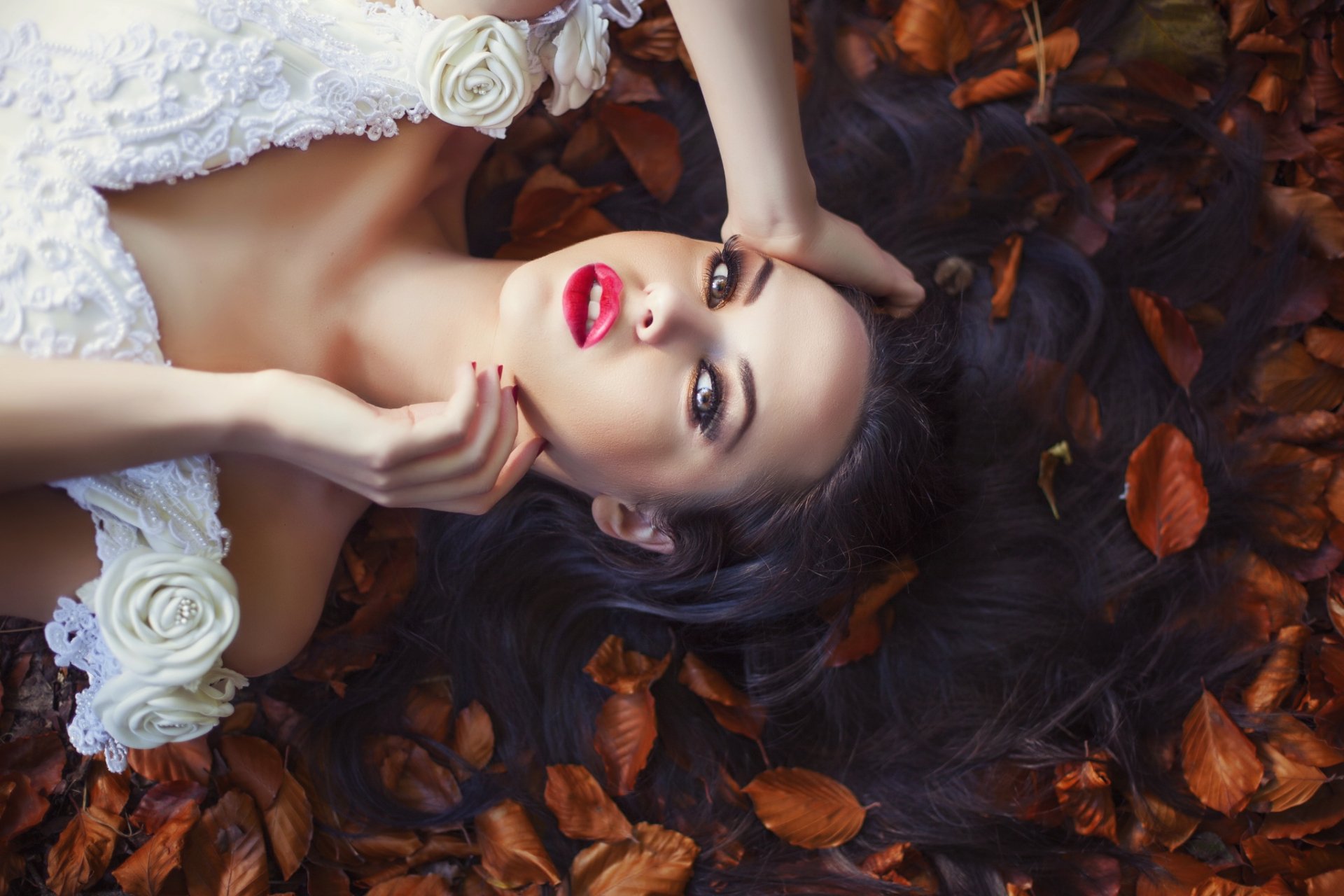A brunette woman with brown eyes lies among brown leaves, wearing a white floral dress, captured in a striking HD desktop wallpaper.