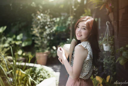 A smiling Taiwanese woman in a polka dot top holds a straw hat while standing in a lush garden, with soft bokeh effects enhancing the serene atmosphere. 4K Ultra HD desktop wallpaper.