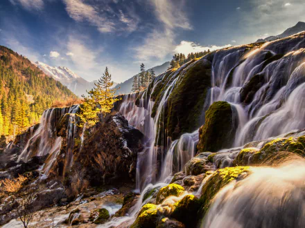 Scenic view of Pearl Shoal Falls cascading over mossy rocks in Jiuzhaigou Park, Sichuan, China, surrounded by mountains under a bright blue sky.