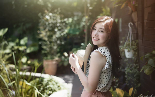 A smiling Taiwanese woman in a polka dot top holds a straw hat while standing in a lush garden, with soft bokeh effects enhancing the serene atmosphere. 4K Ultra HD desktop wallpaper.