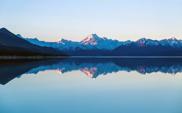 Snow-capped mountains of New Zealand reflect clearly on the calm waters of Lake Pukaki under a bright sky in this 4K Ultra HD nature wallpaper.