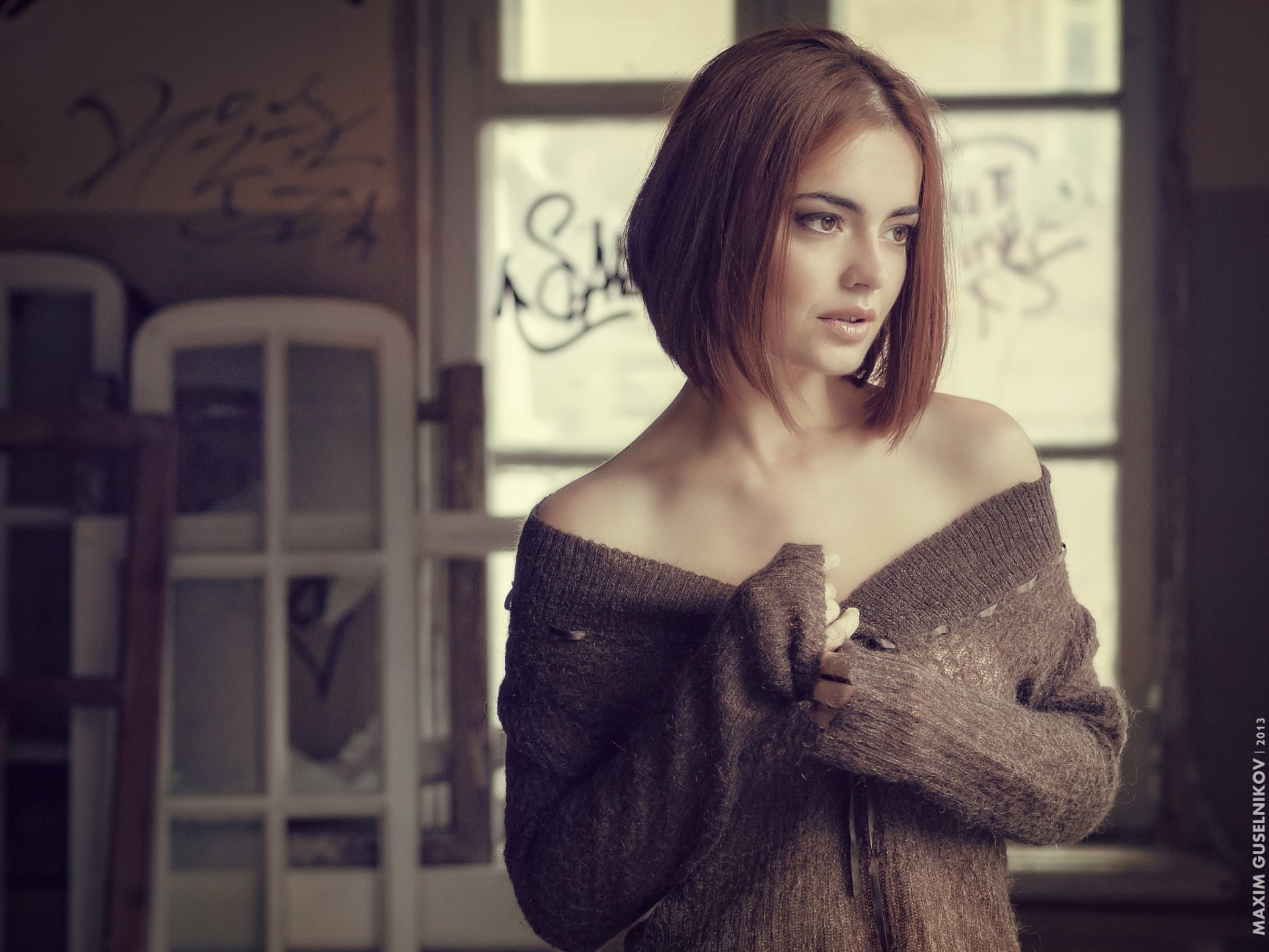 HD desktop wallpaper of a redhead woman with brown eyes, wearing an off-shoulder sweater, posing thoughtfully in a softly lit indoor setting.