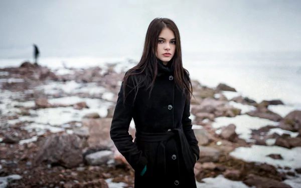A brunette woman with brown eyes stands on a rocky shore, captured with a bokeh effect. This HD desktop wallpaper features model Oktyabrina Maximova.