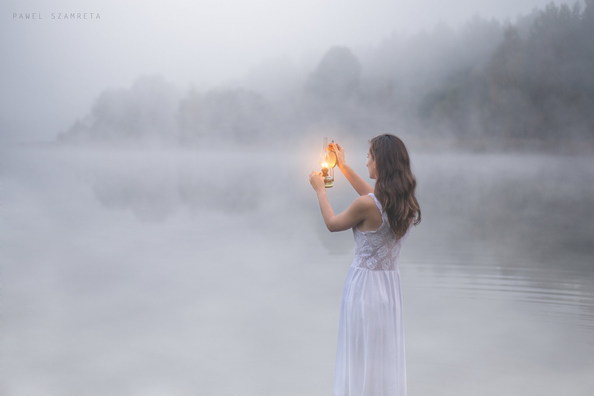 A brunette woman in a dress holds a lantern by a foggy lakeside, creating a moody atmosphere. HD desktop wallpaper.
