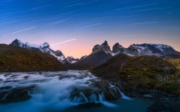 Time-lapse stars streak over the rugged mountains of Torres del Paine, Chile, with a flowing river under a night sky in Patagonia’s stunning natural landscape.
