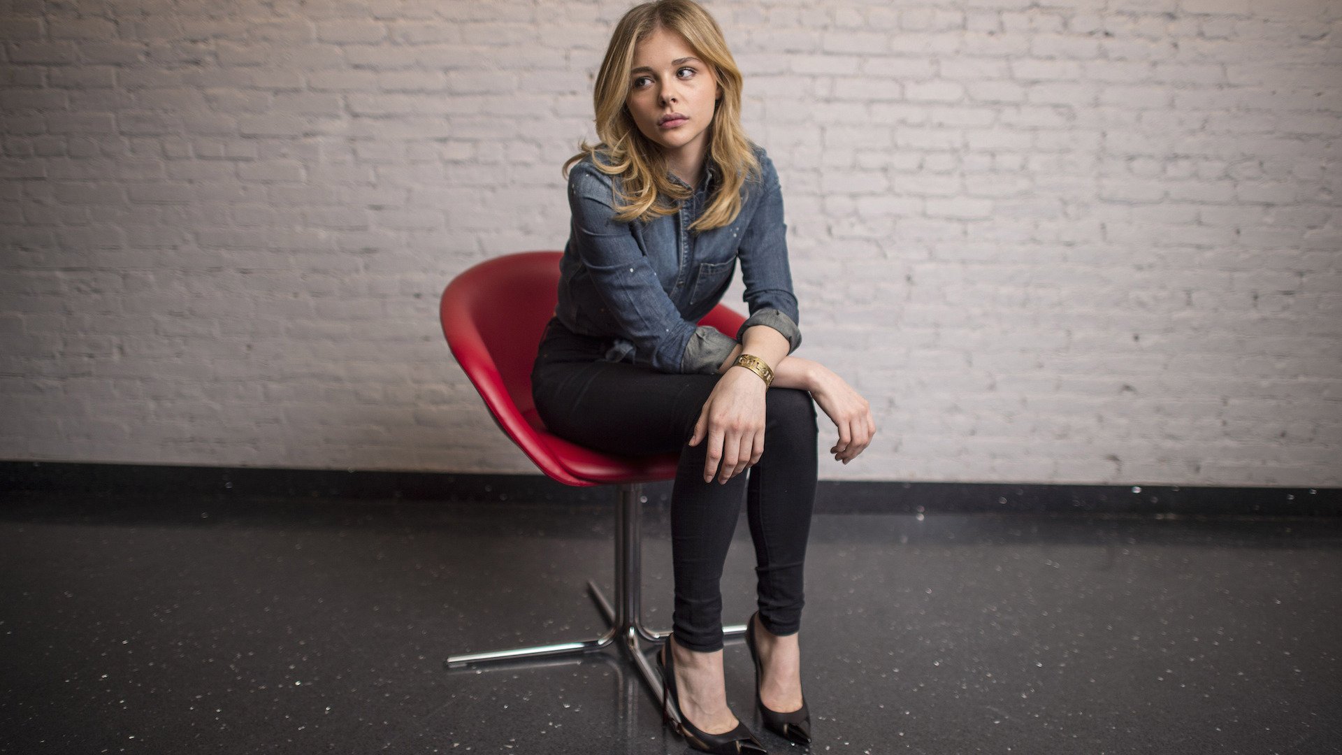 Chloë Grace Moretz poses thoughtfully on a red chair against a textured white wall, captured in this HD desktop wallpaper, showcasing her stylish appearance and expressive gaze.