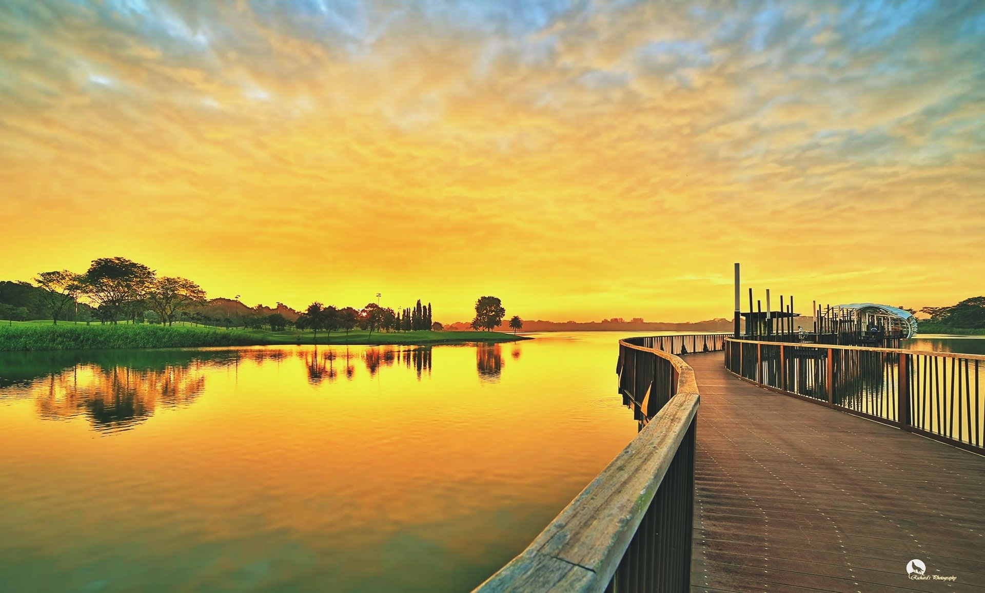 A serene 4K Ultra HD landscape of a wooden bridge over a calm lake, reflecting colorful sky and clouds, surrounded by lush nature and river scenery.