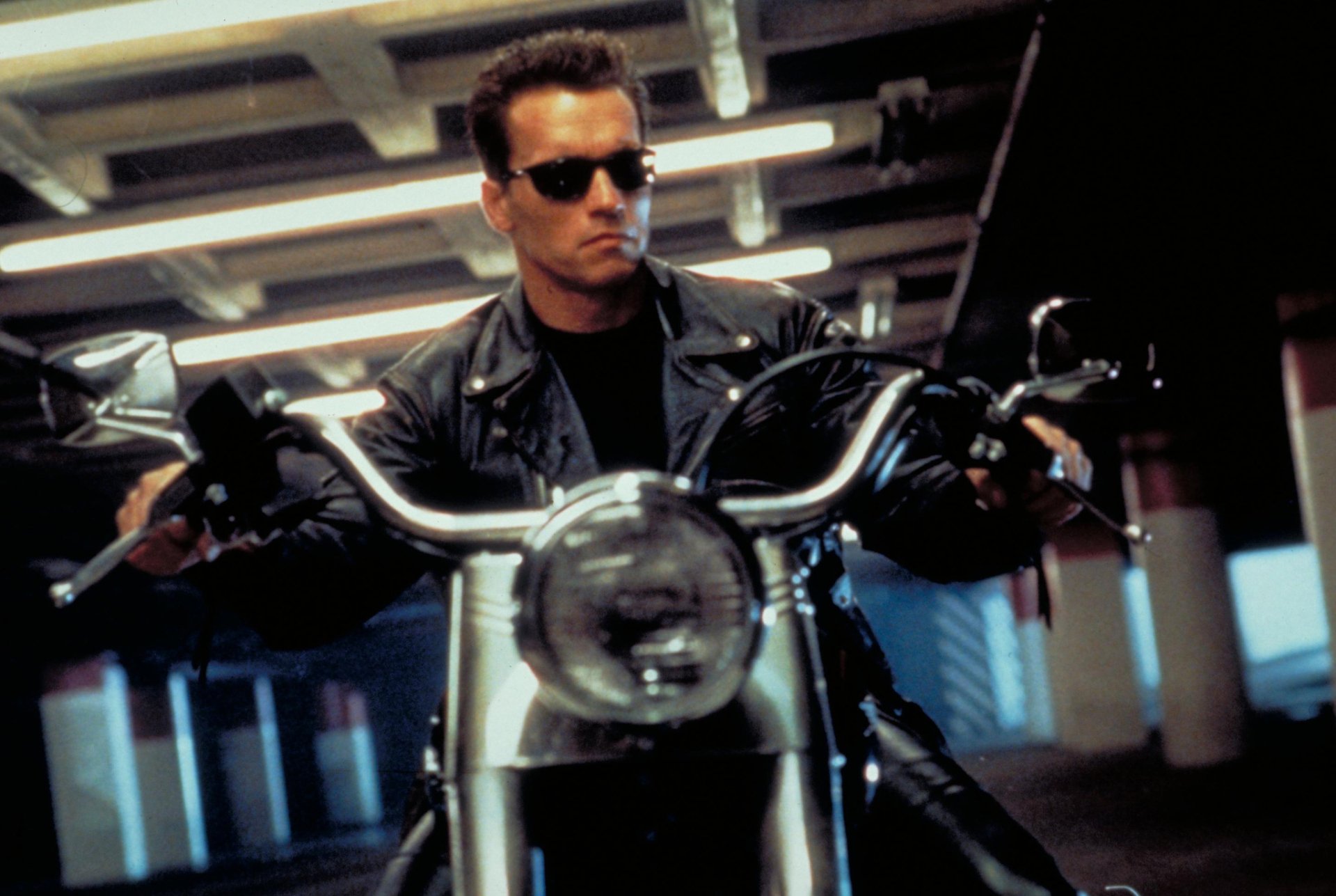 Arnold Schwarzenegger as the Terminator in a sleek black leather jacket, riding a motorcycle, captured in an iconic scene from Terminator 2: Judgment Day. HD desktop wallpaper.
