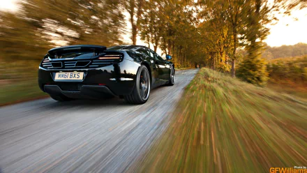 HD desktop wallpaper showcasing a sleek black McLaren MP4-12C speeding down a tree-lined road in autumn.