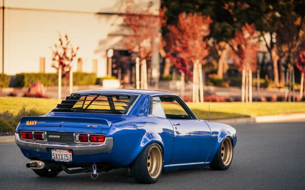 HD PC desktop wallpaper of a blue vintage Toyota Celica parked on a sunlit suburban street, wide wheels and lowered stance, autumn trees and blurred houses in the background.